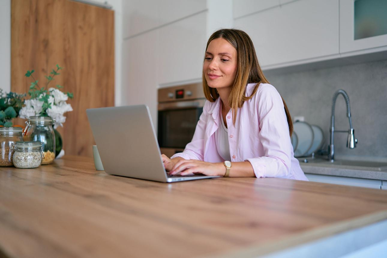Woman in a light pink coat using a laptop at a kitchen counter